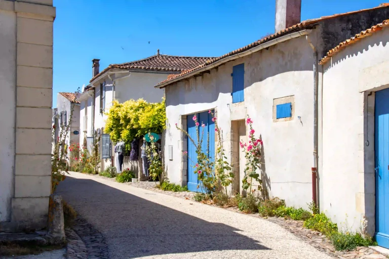 Camping à Talmont-sur-Gironde en Charente-Maritime, ruelle pavée bordée de maisons blanches aux volets bleus et fleurs.